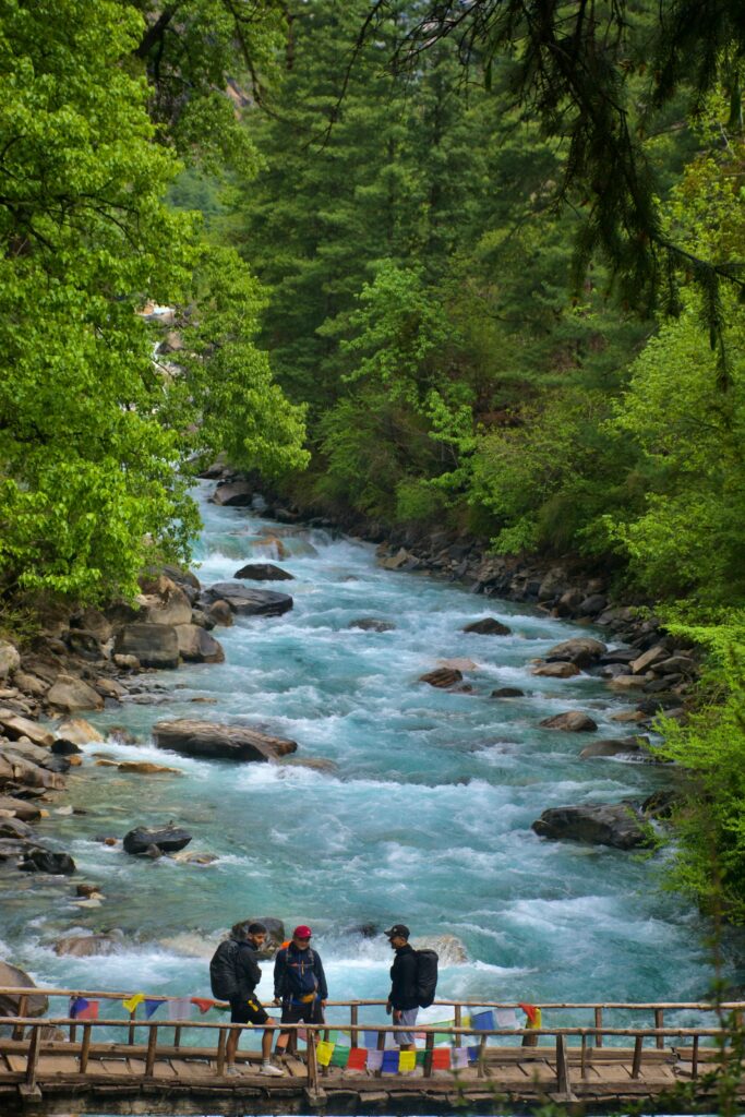 Three hikers standing on a wooden bridge over a wild river surrounded by lush greenery in Nepal.